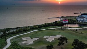 Un campo de golf costero con un faro, un edificio blanco grande y un atardecer sobre el océano.