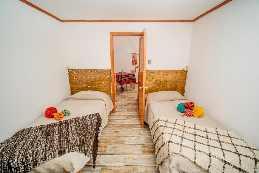 Una habitación pequeña y acogedora con dos camas individuales, cabecero de madera y una mesita con mantel colorido.