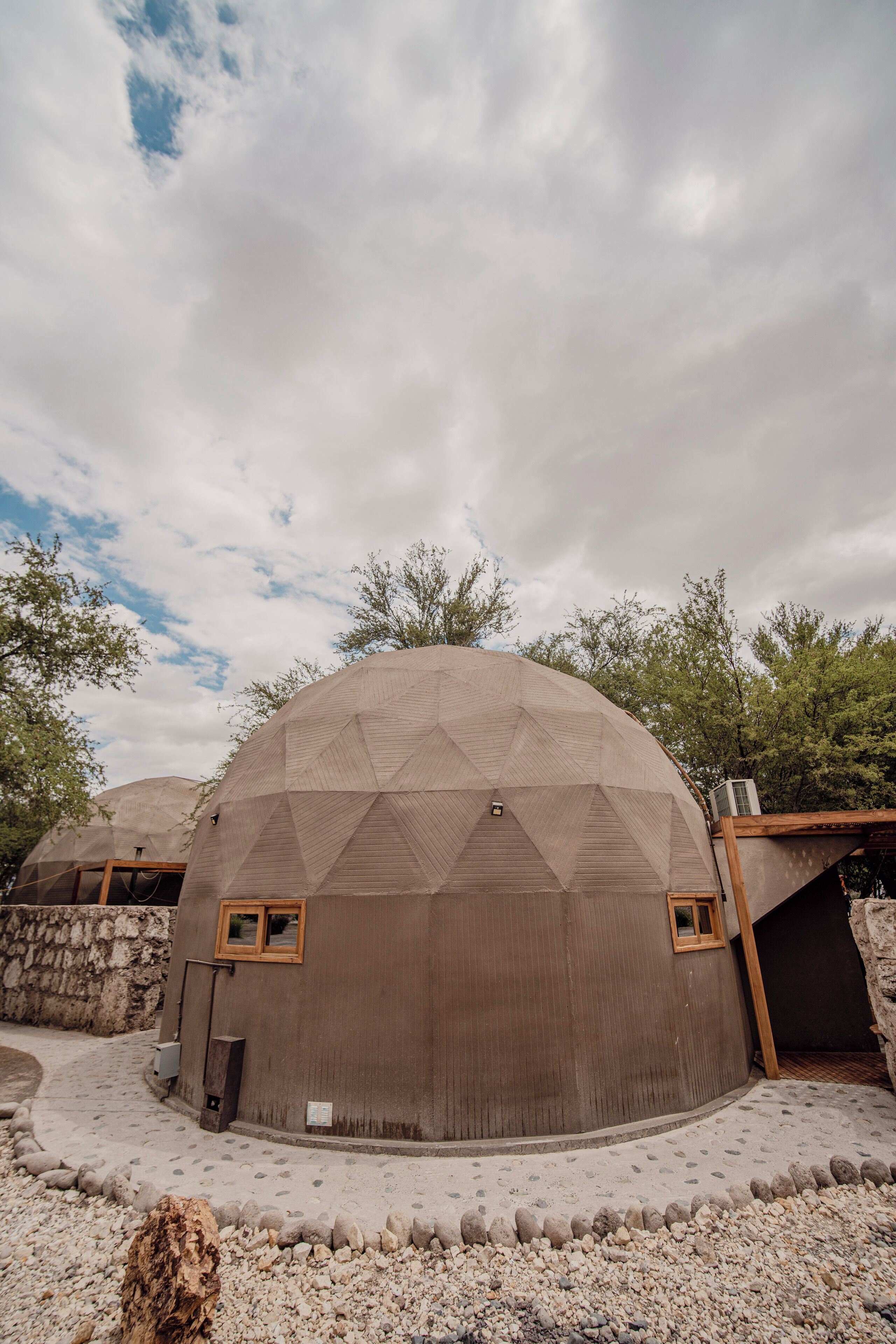 A geodesic dome structure with a window and a door, surrounded by a gravel area and trees.