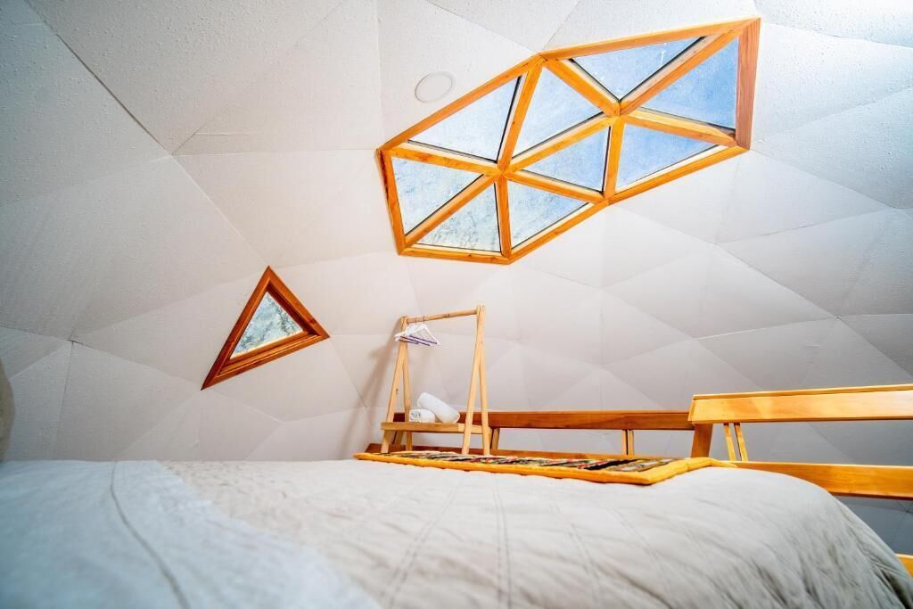 Una habitación con una cama, un banco de madera y una estructura de ventana geométrica.