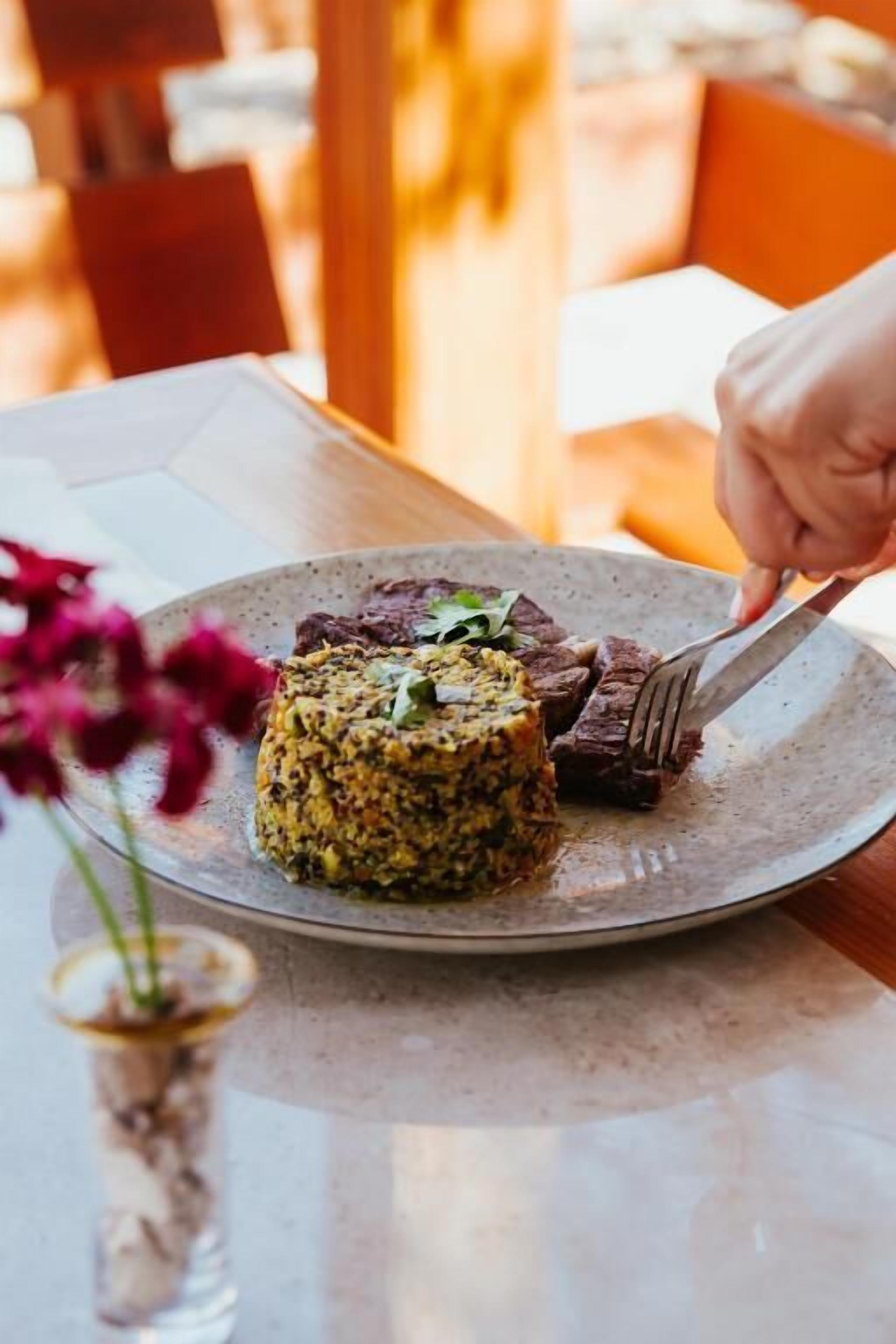Un plato de comida con una mano sosteniendo un tenedor, sobre una mesa con un florero.