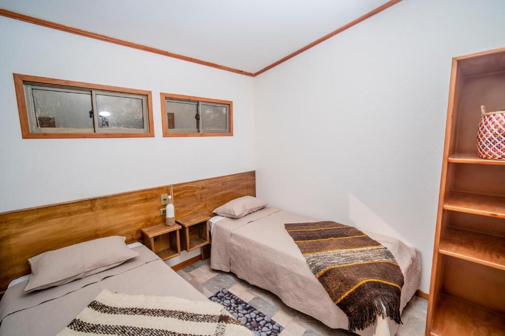 Un dormitorio con dos camas, cabeceros de madera, una mesita de noche, una estantería y dos ventanas.