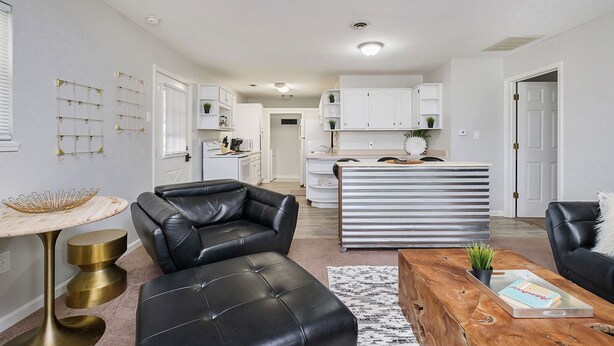 A modern living room with a black leather sofa, a wooden coffee table, and a marble side table. There is a kitchen in the background with white cabinets and a countertop.