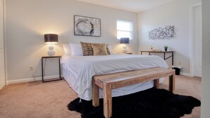 A bedroom with a bed, two bedside tables, a bench, a black furry rug, a window, and abstract wall art.