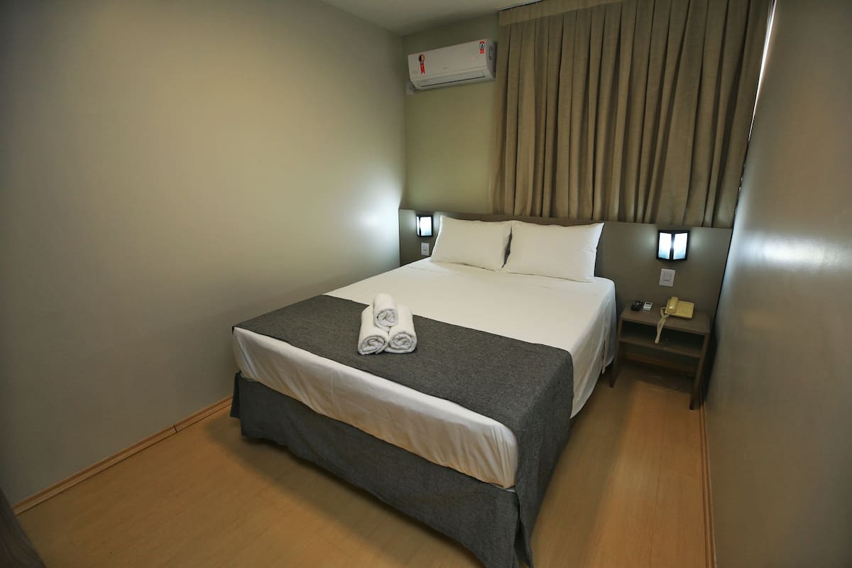 A hotel room with a bed, a bedside table with a lamp, an air conditioning unit, and a curtain.