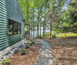 A house with a stone pathway leading to a deck, surrounded by trees and a lake view.