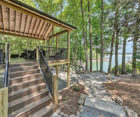 A wooden deck leading to a covered area with outdoor furniture, surrounded by trees and a view of a body of water.