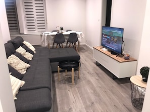 A modern living room with a sectional sofa, a small black ottoman, a dining area with a white table and chairs, and a flat-screen TV on a wooden shelf.