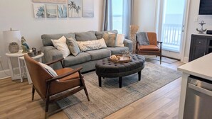 A living room with a grey sofa, brown armchair, ottoman, and a coffee table. There are wall art pieces, a lamp, and a TV mounted on the wall.