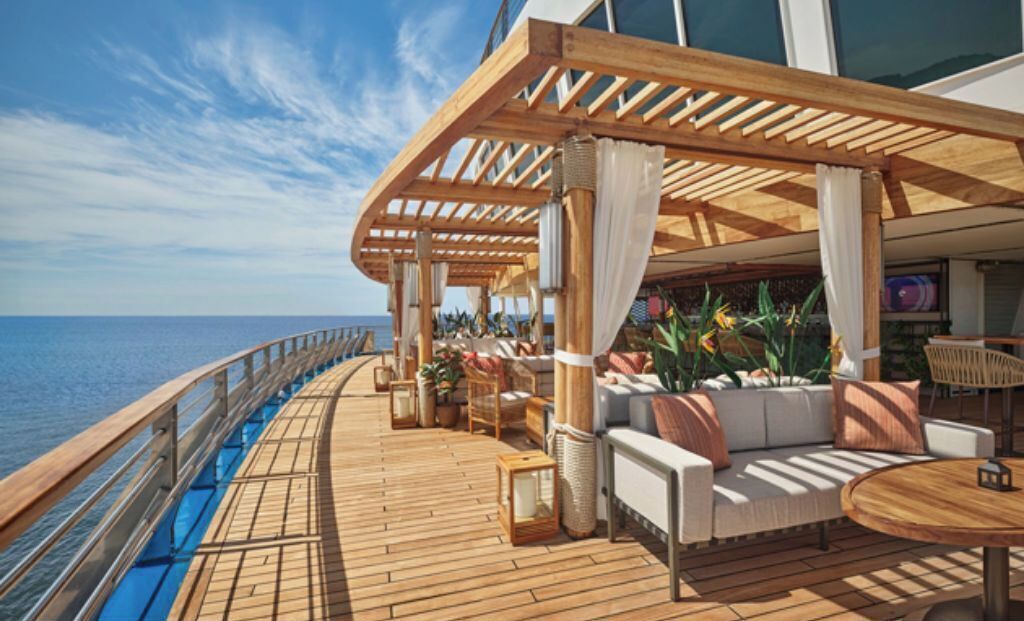Dek van een cruiseschip met houten vloer, een zitgedeelte buiten en uitzicht op de oceaan.