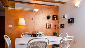 Un comedor con mesa y sillas blancas, aire acondicionado de pared, lámpara, y estantes pequeños con plantas y cuadros.