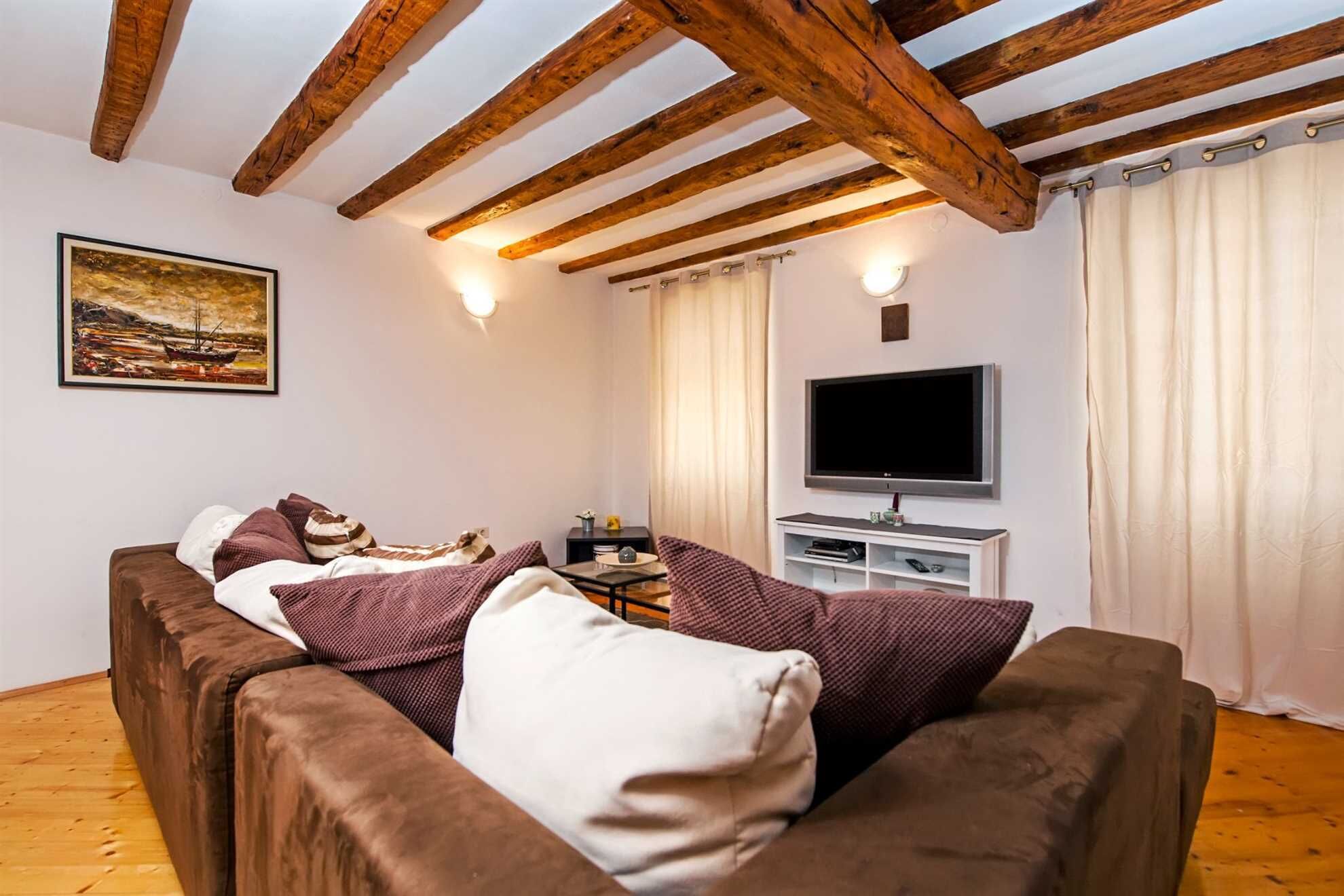 Un salon chaleureux avec un canapé marron, un téléviseur à écran plat, des poutres en bois au plafond et un tableau accroché au mur.