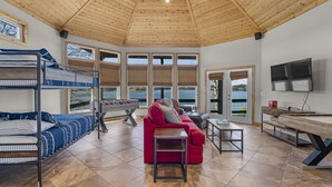 Una habitación amplia con techo alto, literas, un sofá rojo, una mesa de centro, un televisor y grandes ventanales con vista a una masa de agua.