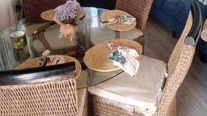 Un coin repas avec une table en verre entourée de chaises en osier, un canapé bleu agrémenté de coussins à motifs et une fenêtre ornée de rideaux légers.