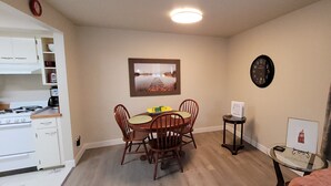 Cocina compacta con electrodomésticos blancos, una mesa de comedor con sillas, un cuadro en la pared, un reloj y un ventanal con cortinas.