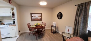 Cocina compacta con electrodomésticos blancos, una mesa de comedor con sillas, un cuadro en la pared, un reloj y un ventanal con cortinas.