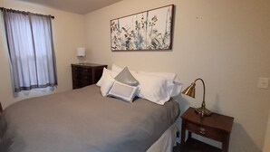 Ein Schlafzimmer mit einem Bett, einem Sessel, einem Nachttisch, einer Lampe, einem Fenster mit Vorhängen und einem Wandbild.