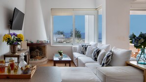 A modern living room with a white sofa, a coffee table, and a TV mounted on the wall. There is a large window with a view of the sea. - Villa Sea Sand - Sunset View Beachfront Villa Near Tavernas & Shops (CHANIA)