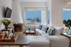Una sala de estar moderna con un sofá blanco, una mesa de centro y un televisor montado en la pared. Hay un ventanal con vistas al mar.