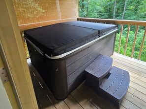 A hot tub with a black cover, a wooden deck, and a view of a forest. - Firefly Landing at Avenwood Cabins (New Straitsville)