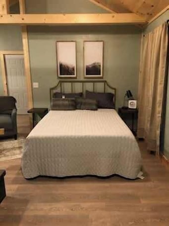 A bedroom with a bed, two framed pictures on the wall, a nightstand, and a chair.. Firefly Landing at Avenwood Cabins