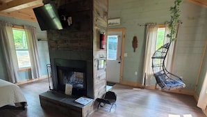 A spacious room with a fireplace, wooden floors, a hanging chair, and a television. - Firefly Landing at Avenwood Cabins (New Straitsville)