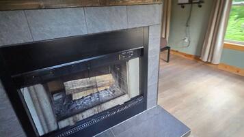A fireplace with a stone surround, a wooden mantel, and a view of a green outdoor area through a window.
