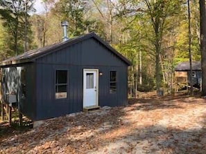 A small, dark-colored cabin with a white door, surrounded by trees and a dirt path.