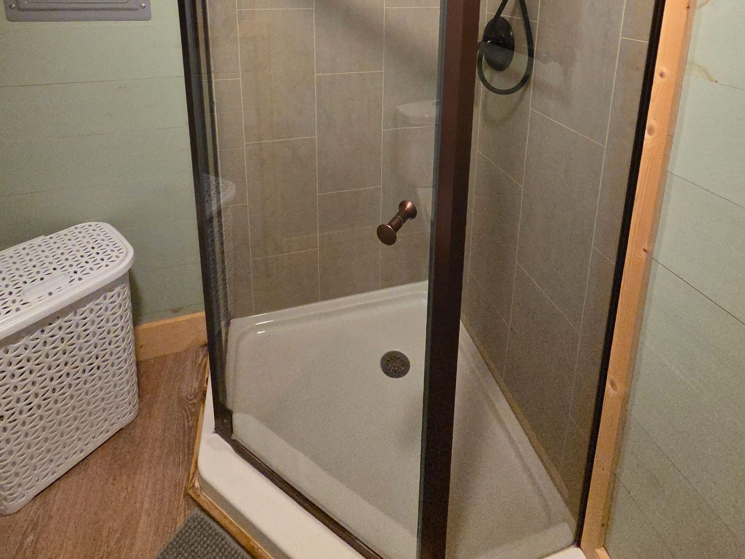 Área de chuveiro com porta de vidro, chuveiro e cesta de plástico branco.