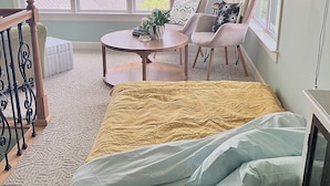 Ein Zimmer mit einem Bett, zwei Stühlen, einem kleinen Tisch und Blick auf ein Gewässer durch große Fenster.