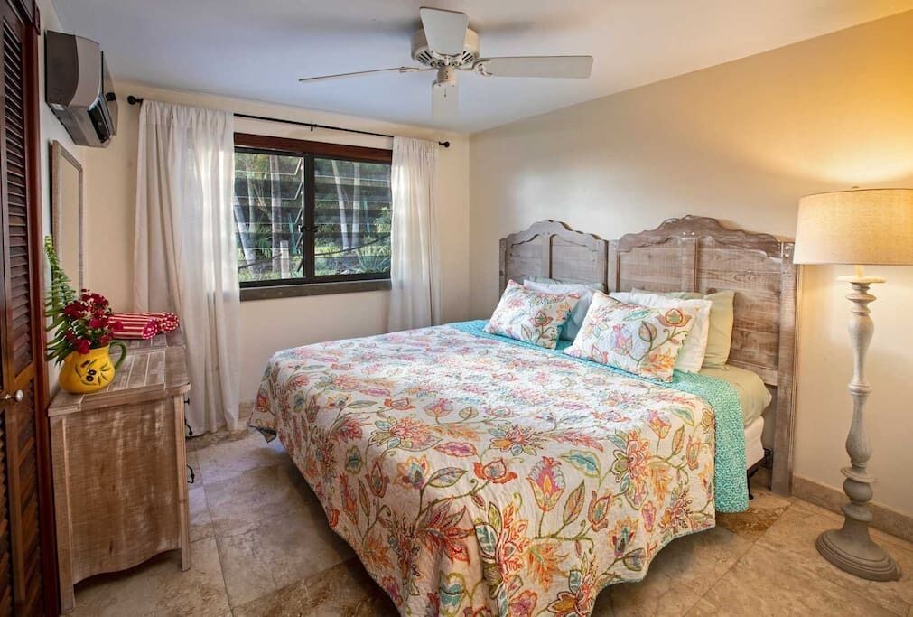 Ein Schlafzimmer mit Bett, Deckenventilator, Fenster mit Vorhängen, Nachttischlampe und einem kleinen Tisch mit Vase und Blumen.