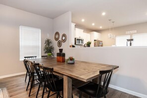 A dining area with a wooden table and black chairs, a kitchen in the background, and a potted plant on the table.