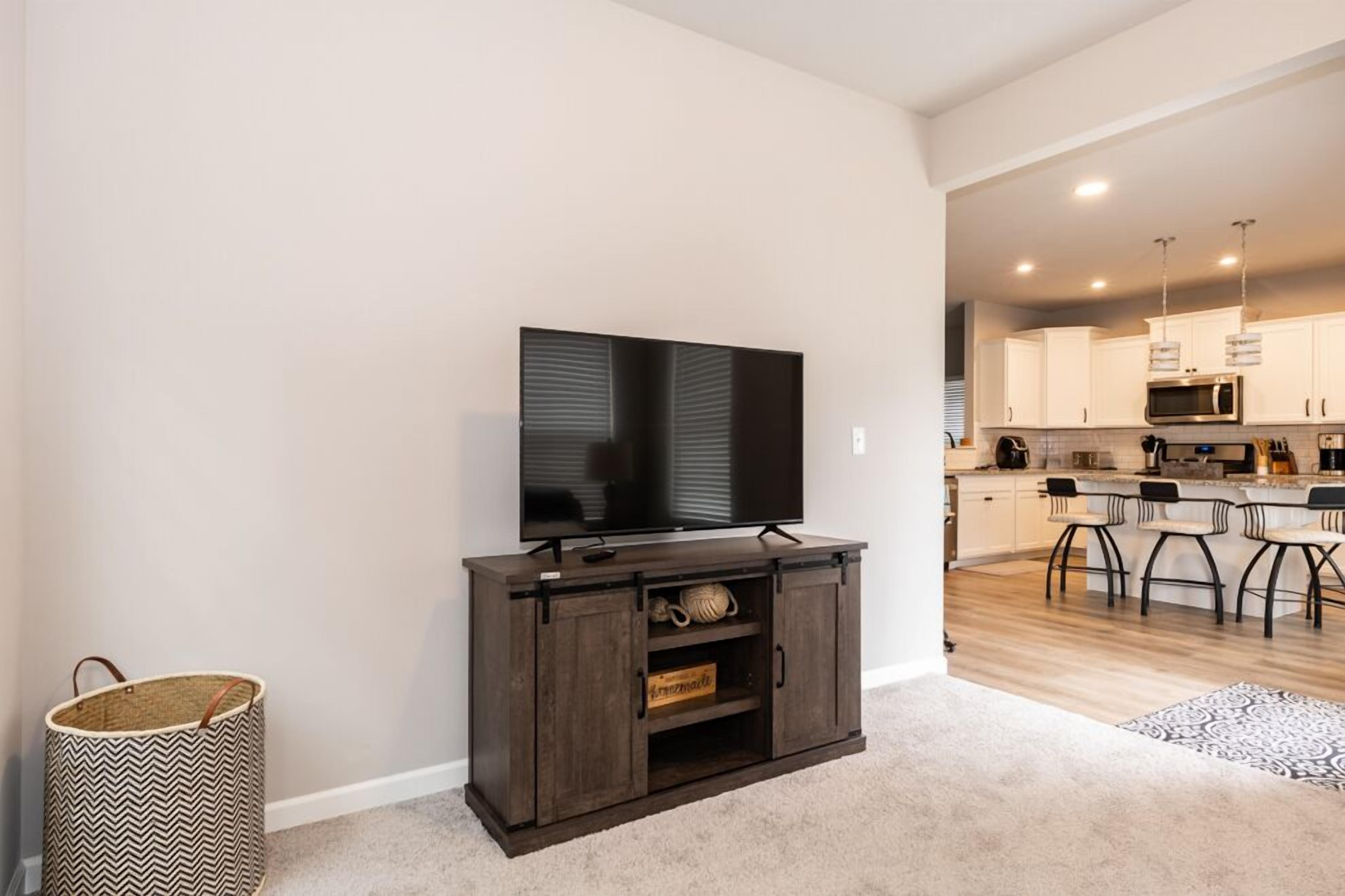 A modern kitchen with a flat-screen TV on a wooden cabinet, a kitchen island with bar stools, and a visible trash bin.