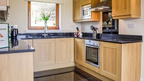 A modern kitchen with wooden cabinets, a black countertop, a built-in oven, and a window with a view of greenery. - 2 bedroom accommodation in Blairgowrie (Blairgowrie)