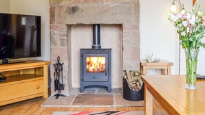 A stone fireplace with a modern wood-burning stove, a flat-screen TV on a wooden stand, a vase with flowers, and a wooden table. - 2 bedroom accommodation in Blairgowrie (Blairgowrie)