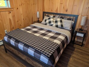 Una cama con colcha de cuadros y varias almohadas, una mesita de noche con lámpara y una pared de madera.