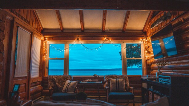L’intérieur chaleureux d’un chalet avec un plafond en bois, une table entourée de chaises, un téléviseur et une vue sur l’eau.