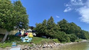 Un lac aux eaux limpides, bordé d’une rive rocheuse, une zone herbeuse avec des radeaux gonflables, et une maison surmontée d’un parasol bleu.