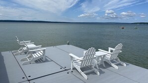 Une terrasse avec des chaises Adirondack blanches et une table, donnant sur un lac paisible où l’on aperçoit des bateaux et des collines au loin.