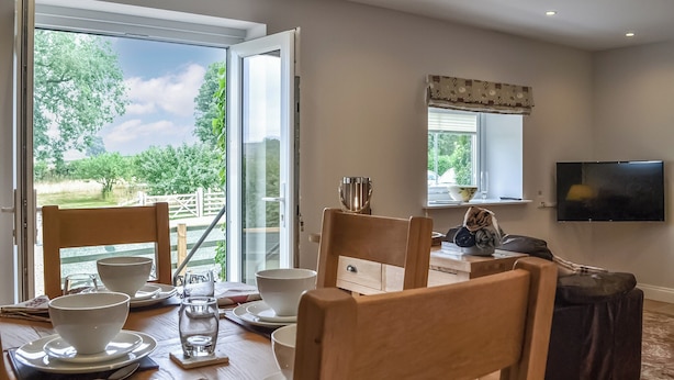 Ein Essbereich mit einem Tisch für vier Personen, einem an der Wand befestigten Fernseher und einem Fenster mit Blick auf Grünflächen.