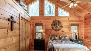 A wooden cabin bedroom with a bed, two nightstands, a cross on the wall, and a ceiling fan. - Expansive Lake Front Log Cabin/Easy Walk to Private! (Jacksboro)