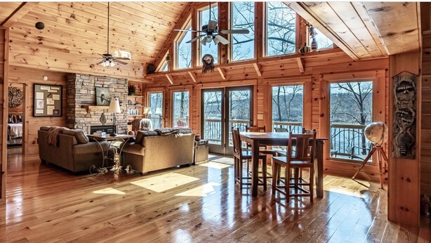 A spacious cabin interior with a wooden floor, a dining area with a table and chairs, a fireplace, and large windows offering views of the outdoors. - Expansive Lake Front Log Cabin/Easy Walk to Private! (Jacksboro)