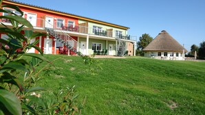A multi-story building with a red and yellow facade, a thatched-roof structure, and a grassy lawn. - Vacation resort Süderhof (Breege)