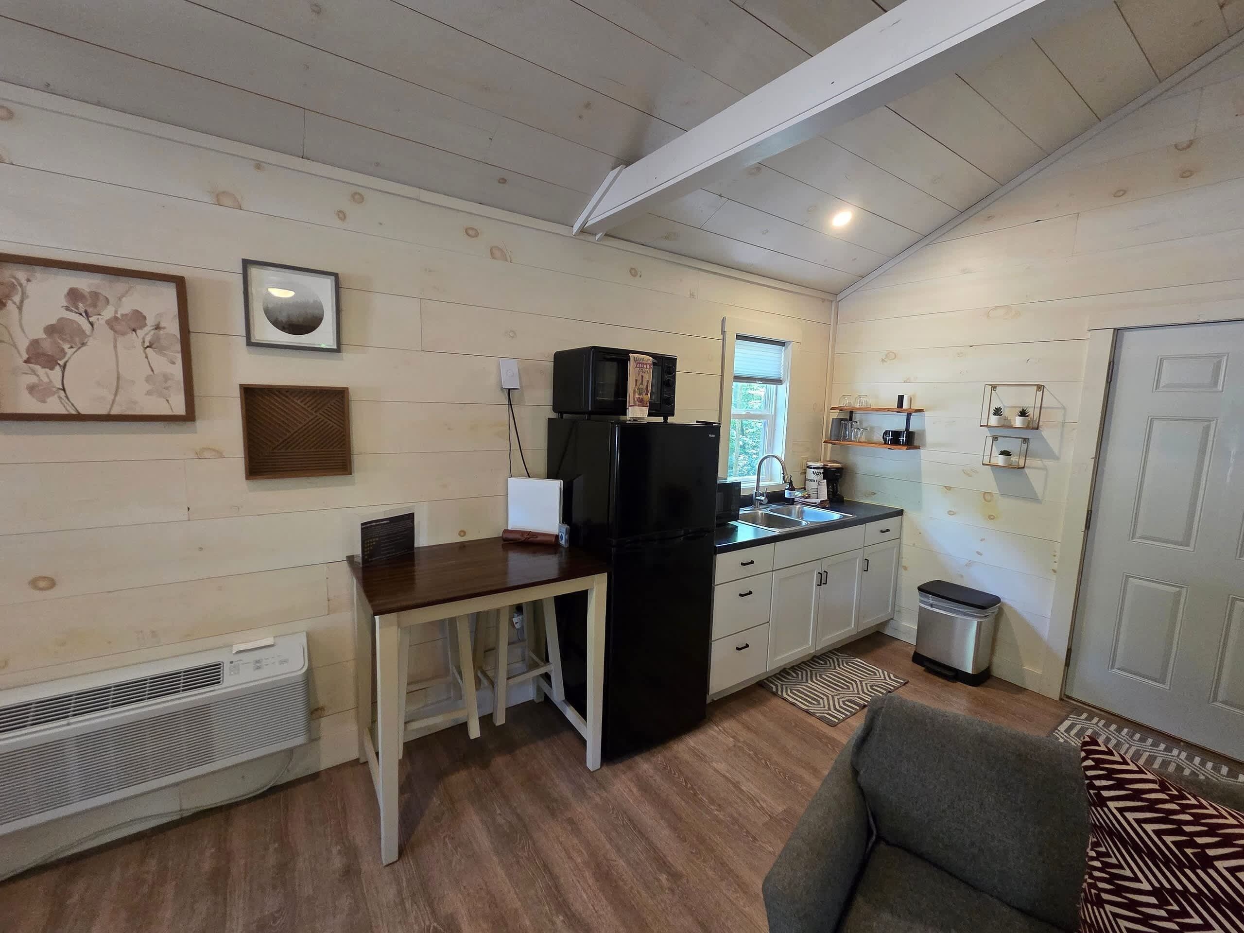 A compact kitchen with a refrigerator, microwave, and sink. There is a dining table with chairs, a small trash bin, and a window with blinds.