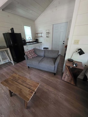 Un espace de vie compact comprenant un canapé, une table basse en bois, un coin cuisine avec un réfrigérateur et une porte donnant sur une autre pièce.
