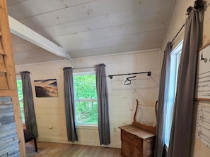 A cabin interior with a wooden dresser, a window with curtains, a painting on the wall, and a bulletin board with text.