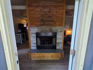 A fireplace with a stone surround and a wooden mantel, located in a room with wooden flooring and a door frame visible. - Owl's Tale at Avenwood Cabins (New Straitsville)