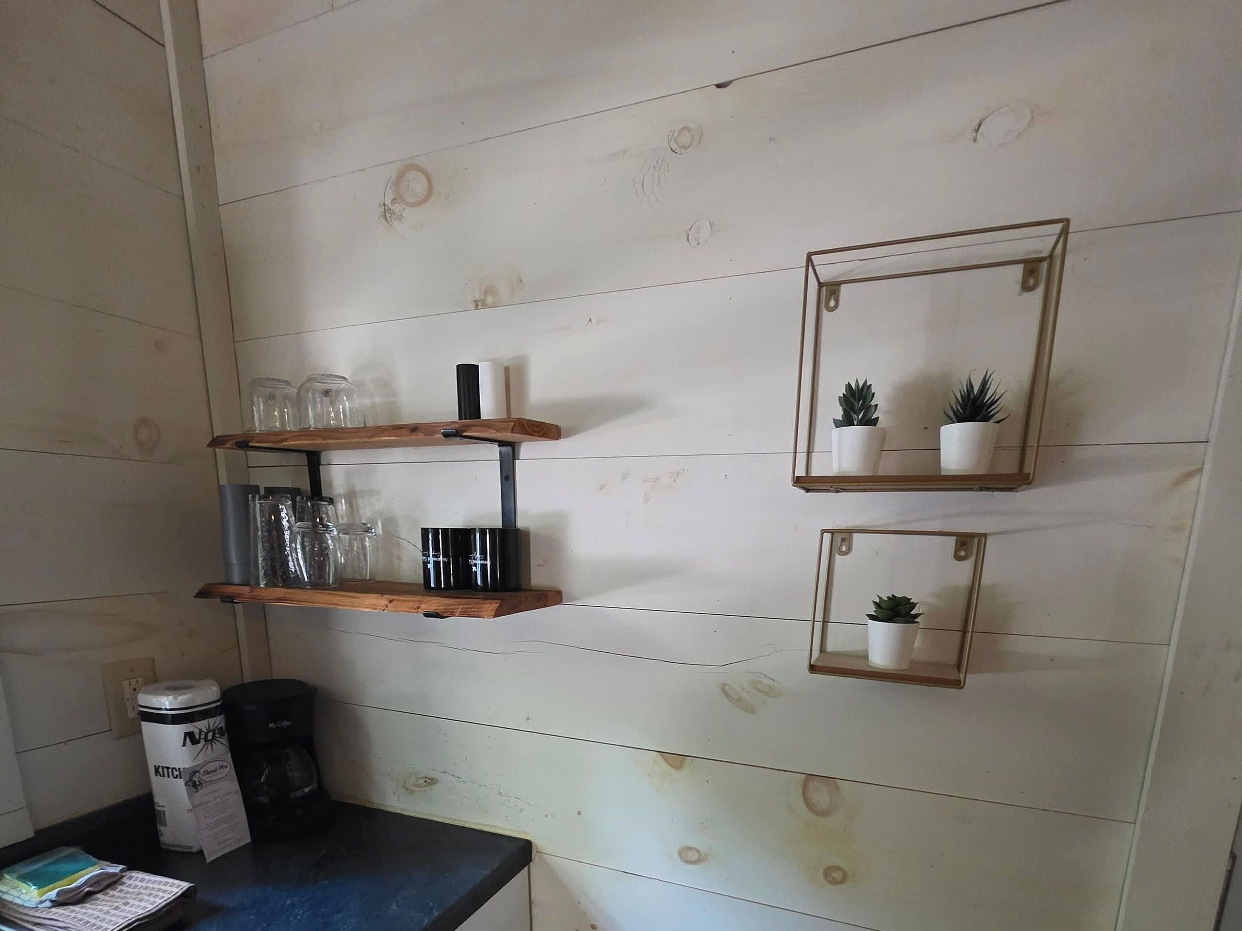 Uma cozinha com prateleiras de madeira, uma cafeteira e dois espelhos fixados na parede com plantas em vasos.