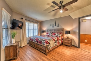 A bedroom with a bed, wooden floor, ceiling fan, wall-mounted TV, bedside tables with lamps, and a window with blinds. - Gatlinburg Cabin  Hot Tub Fire Pit & Amazing Views (Gatlinburg)