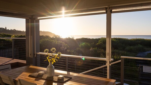 Un balcon avec une table en bois, un vase rempli de fleurs et une vue sur l’océan et la végétation.